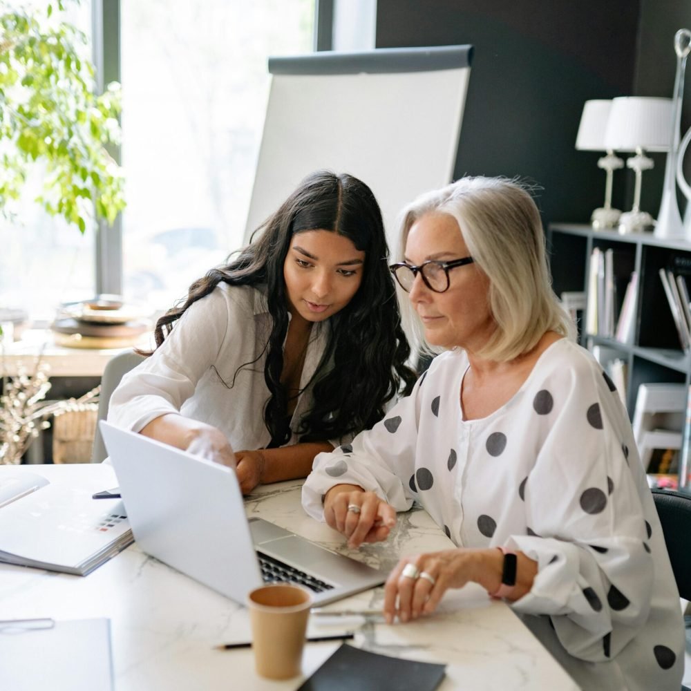 Two professional women collaborating on a project with a laptop in a modern office setting.